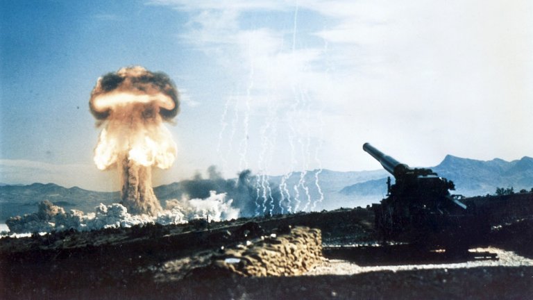 A howitzer sits on the foreground while a mushroom cloud detonates in the background.