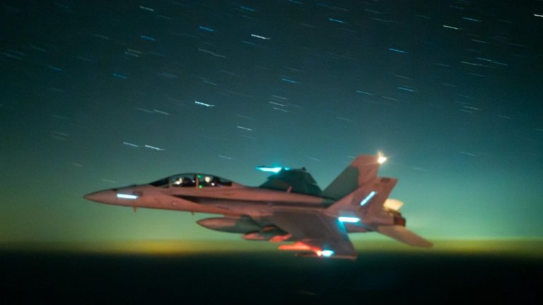 A F/A-18 Super Hornet flies in the night sky, with several stars visible behind it.