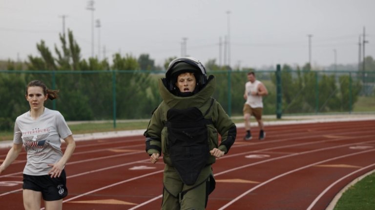 A sweating man in a bulky black and green bomb protection suit runs on a track.