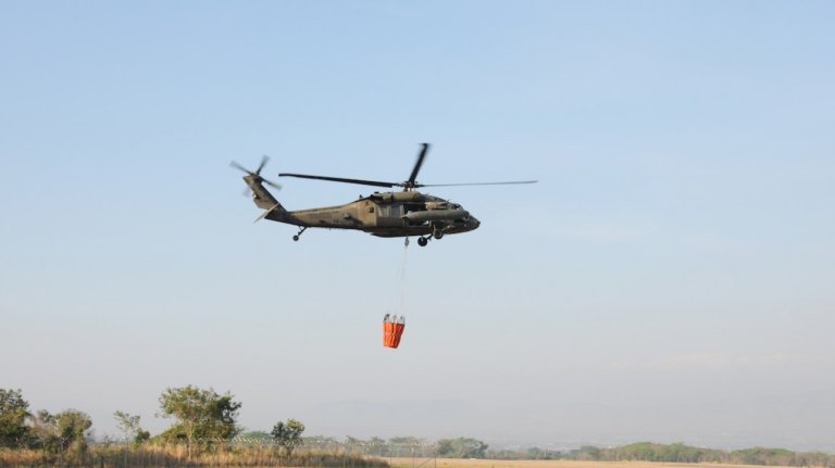 A helicopter carries a bucket over a desert terrain.