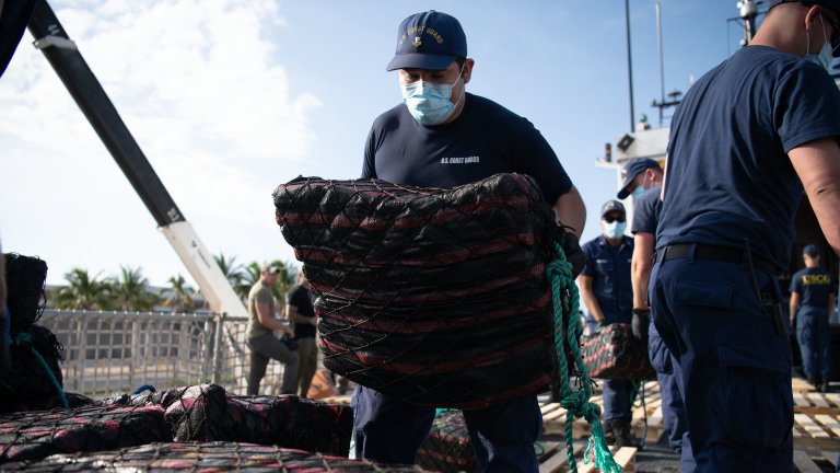 Several Coast Guard crewmembers in face masks move wrapped bundles of drugs.